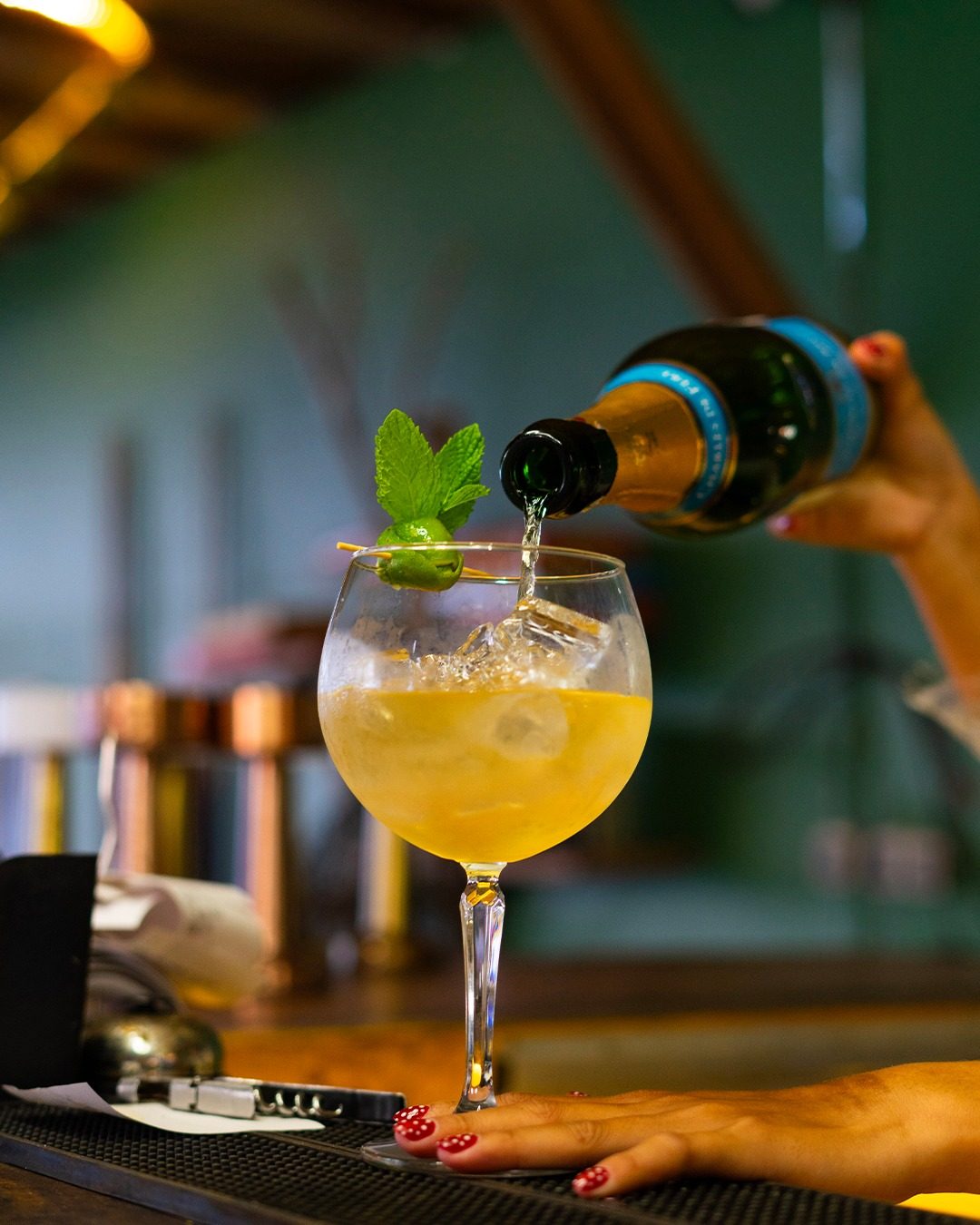 A bartender pouring sparkling wine into a gin glass with mint, bar lights behind
